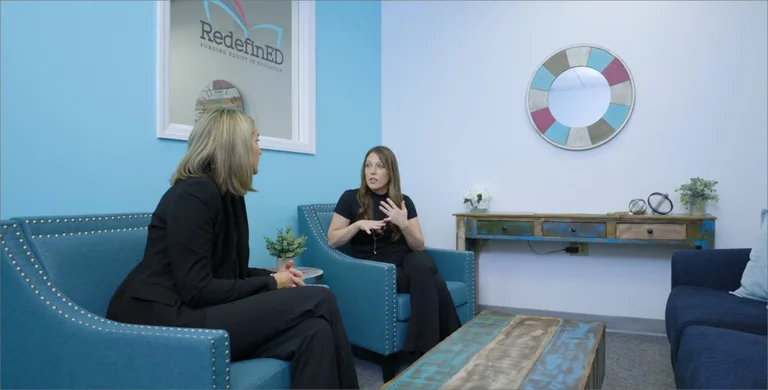 Two women sit facing each other in a light blue room, appearing to converse.
