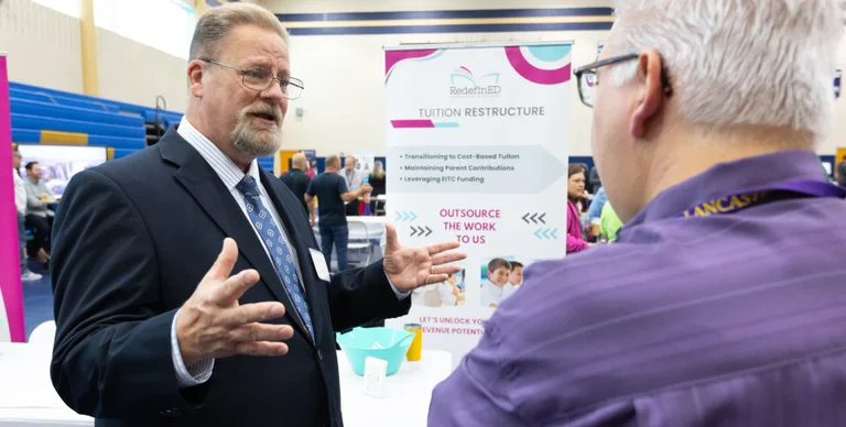 A man in a suit gestures while speaking with another person at a conference booth.