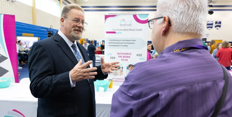 A man in a suit speaks with another attendee at a conference about tuition restructure.