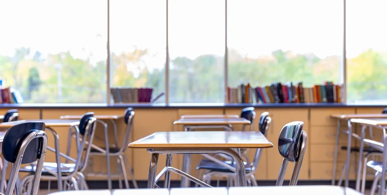 Empty school desks and chairs sit in front of a large window showcasing trees.
