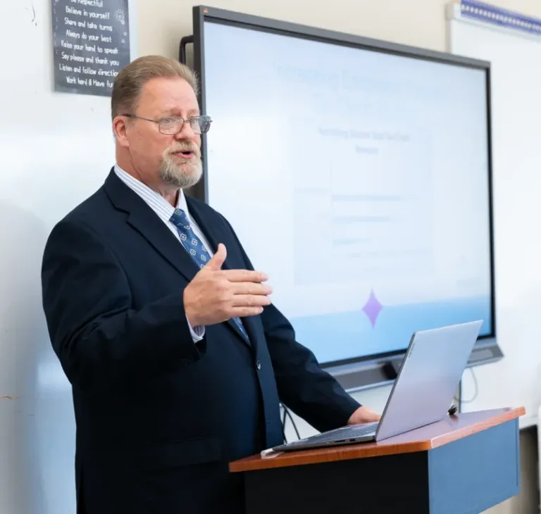 A man with glasses and a beard gestures while speaking at a podium with a laptop.