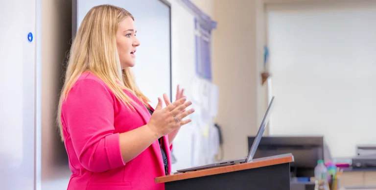A woman in a pink blazer gestures while speaking at a podium with a laptop.