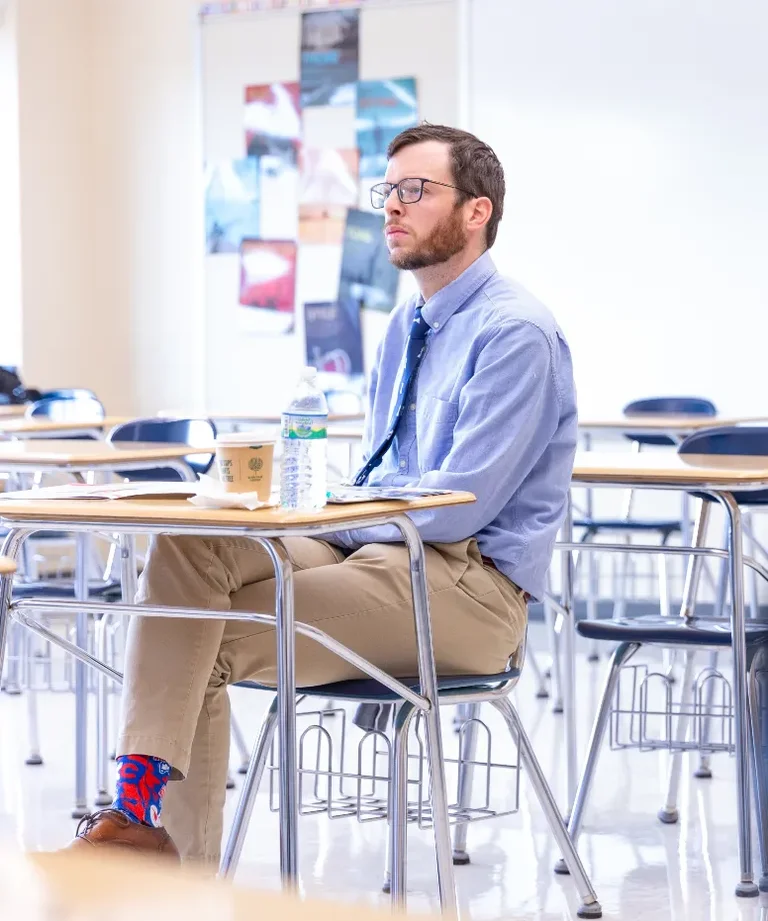 A man in a blue shirt and khaki pants sits at a desk in a classroom.