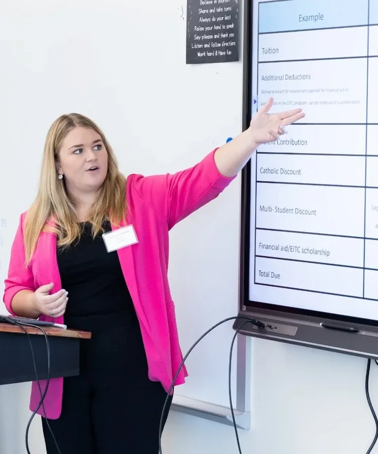 A woman in a pink blazer points to a whiteboard with financial aid information during a presentation.