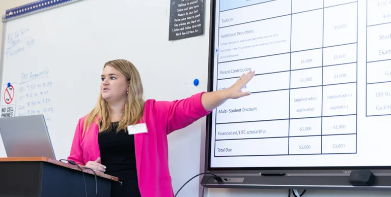 A woman in a pink blazer gestures toward a whiteboard listing financial aid and tuition information.