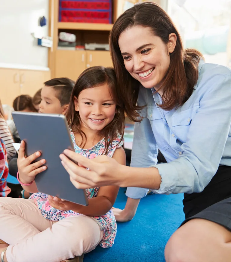 A smiling teacher sits with a young girl looking at a tablet together in a classroom.