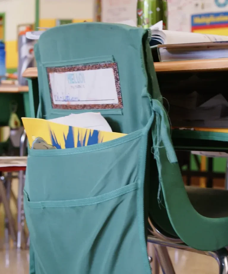 A green chair caddy hangs over a classroom chair, holding papers and a name tag labeled 'Helion.'