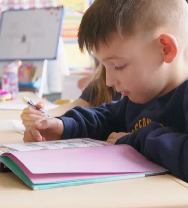 A young boy concentrates while coloring in a book at a desk.