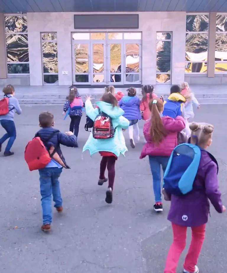 Children with backpacks run excitedly outside a building towards the street.