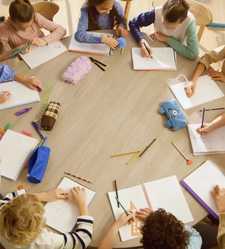 Several children sit around a table, drawing and writing with colored pencils and pens.