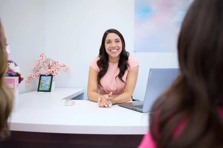 A smiling woman with brown hair sits at a desk, looking toward the viewer and gesturing with her hands.