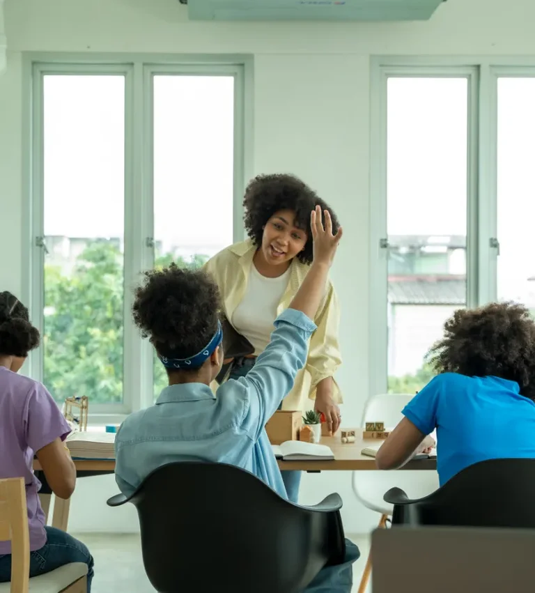 A Black woman raises her hand while speaking to three students seated at a table in a classroom.