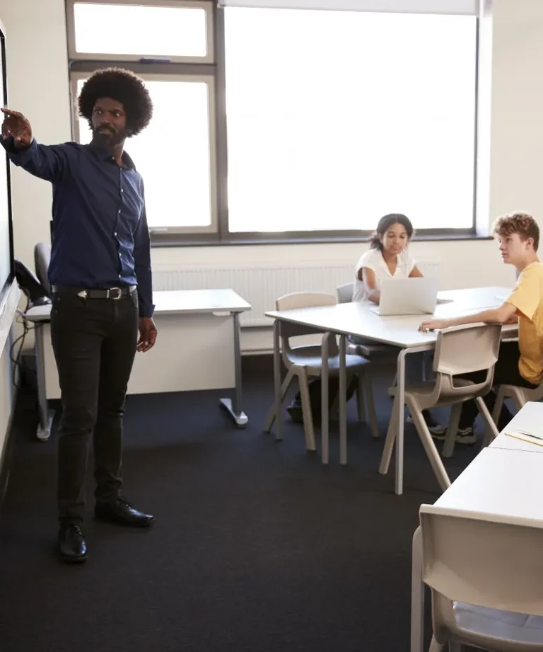 A man in a dark shirt points toward a whiteboard while two students sit at desks in a classroom.