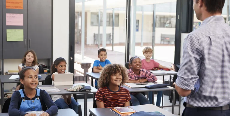 A teacher lectures to a group of elementary school students seated at desks in a classroom.