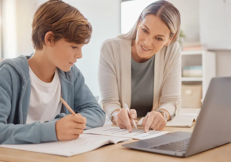 A woman assists a young boy with schoolwork at a table with a laptop and papers.