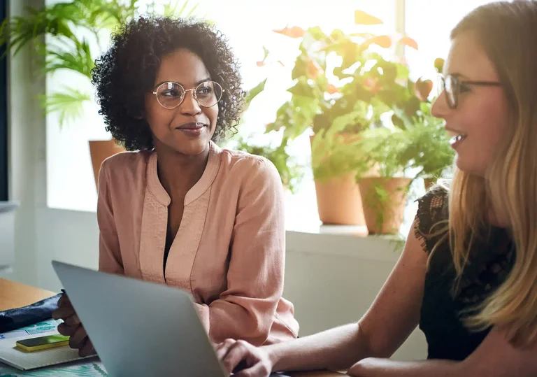 Two women with glasses sit at a table, smiling and conversing while looking at a laptop.