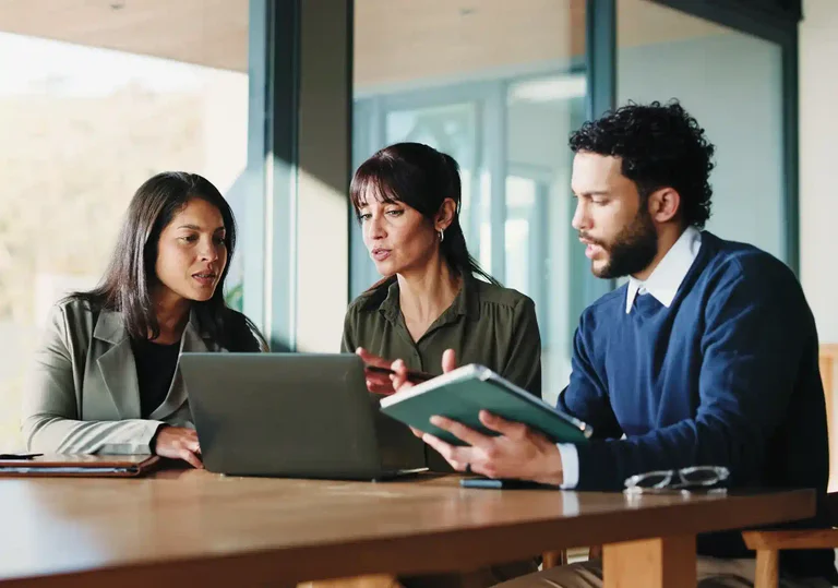 Three colleagues discuss work on laptops and tablets at a modern wooden table.